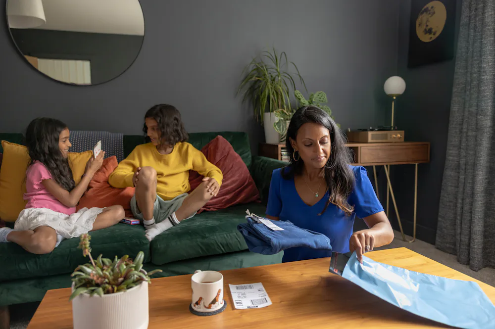 A mother is packaging a pair of trousers into a parcel bag with the delivery label ready to be attached. Her two children are sat playing cards on the sofa behind her