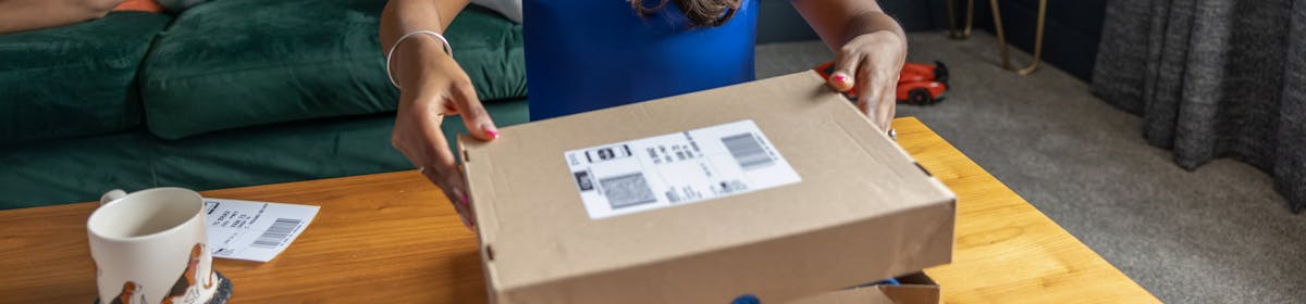A mother is placing the lid on a parcel box with the shipping label attached so it is ready to send. Her two children are sat on the sofa behind her reading a book.