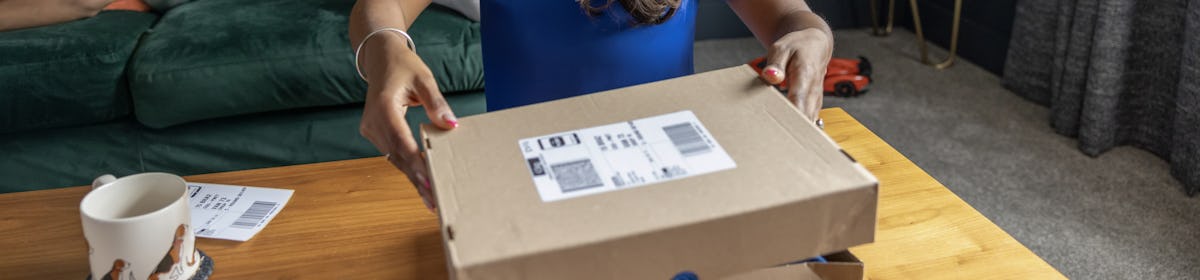 A mother is placing the lid on a parcel box with the shipping label attached so it is ready to send. Her two children are sat on the sofa behind her reading a book.