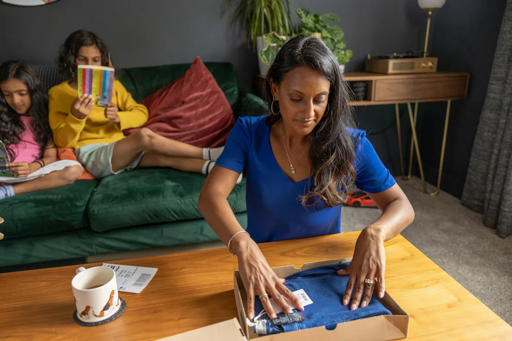 A mother is placing a pair of jeans into a parcel box with the shipping label sat beside ready to attach. Her to children are reading a book on the sofa behind her.