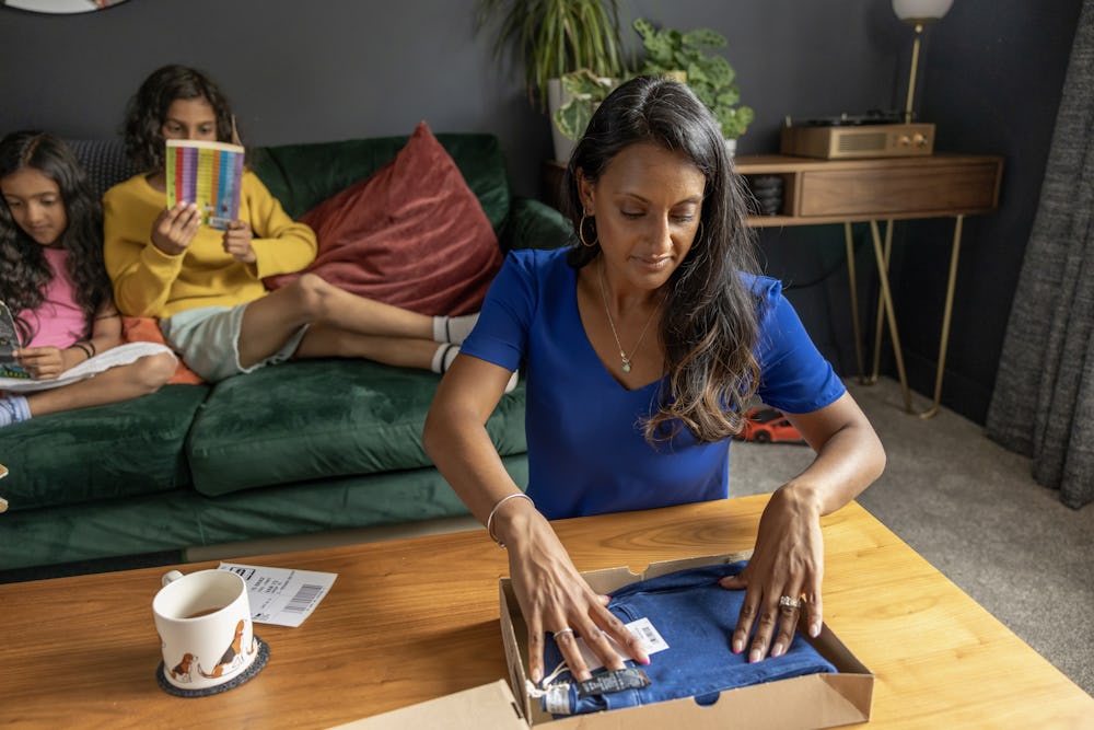 A mother is placing a pair of jeans into a parcel box with the shipping label sat beside ready to attach. Her to children are reading a book on the sofa behind her.