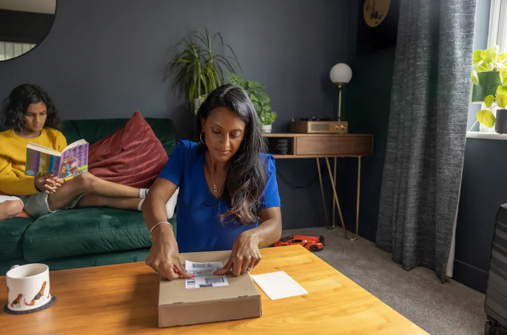 A mother is attaching the shipping label to a parcel on her coffee table. Her two children are sat behind her reading a book.