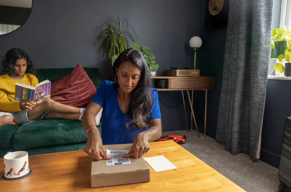 A mother is attaching the shipping label to a parcel on her coffee table. Her two children are sat behind her reading a book.