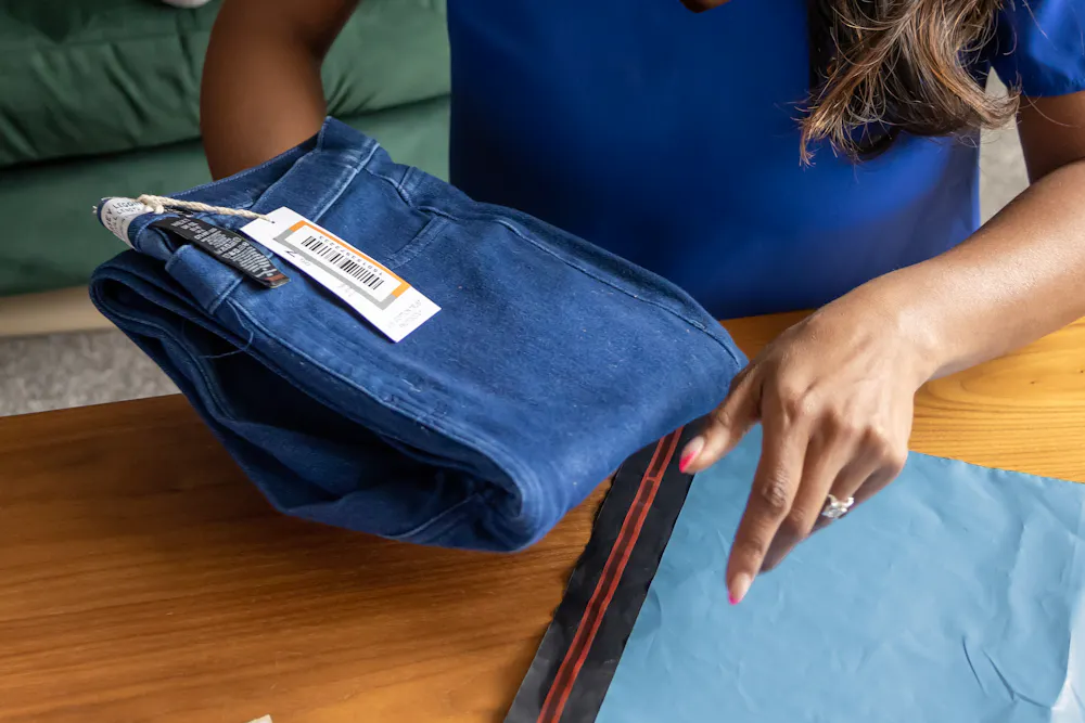 A mother is placing a pair of jeans into a parcel bag with the shipping label sat beside ready to attach. Her to children are watching a tablet on the sofa behind her.