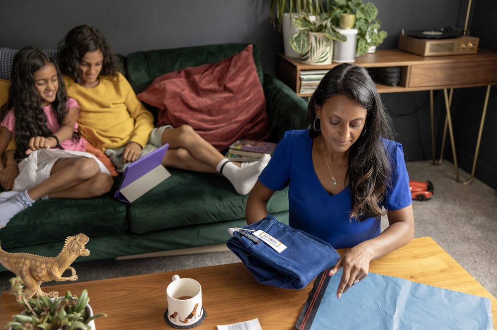 A mother is placing a pair of jeans into a parcel bag with the shipping label sat beside ready to attach. Her to children are watching a tablet on the sofa behind her.