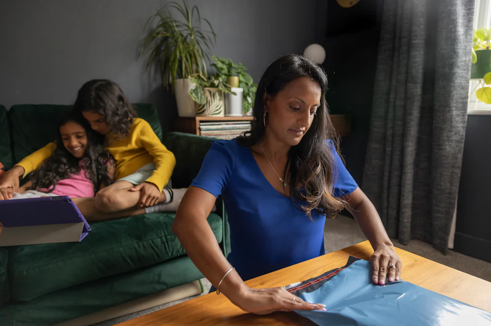 A mother is sealing a parcel bag on her coffee table. Her two children are watching a tablet on the sofa behind her