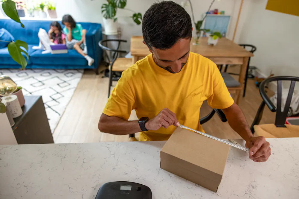 A father is measuring the sides of his parcel at home at his desk using measuring tape. His children are resting on the sofa behind him