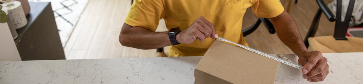 A father is measuring the sides of his parcel at home at his desk using measuring tape. His children are resting on the sofa behind him