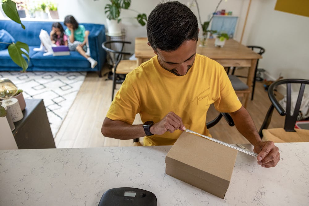 A father is measuring the sides of his parcel at home at his desk using measuring tape. His children are resting on the sofa behind him