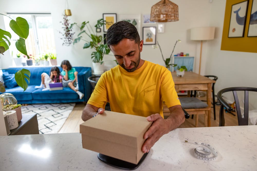 A father is weighing his parcel at home at his desk using a set of digital scales. His children are resting on the sofa behind him.