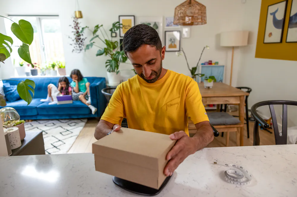 A father is weighing his parcel at home at his desk using a set of digital scales. His children are resting on the sofa behind him.