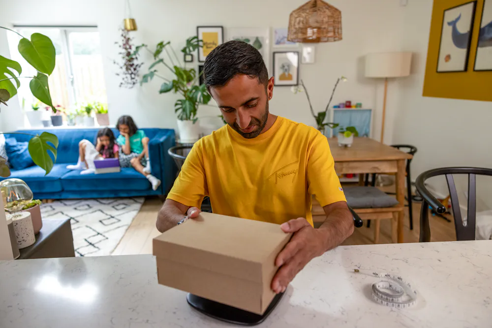 A father is weighing his parcel at home at his desk using a set of digital scales. His children are resting on the sofa behind him.