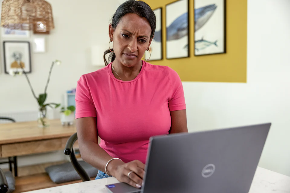 A woman is sat at home at her desk looking puzzled or confused towards her laptop screen