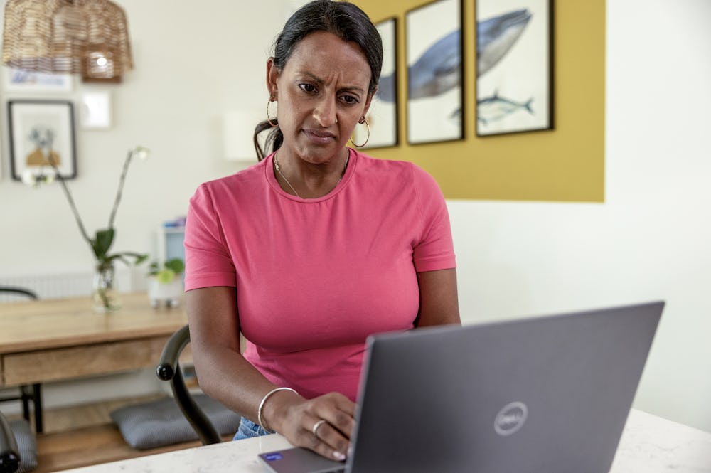 A woman is sat at home at her desk looking puzzled or confused towards her laptop screen