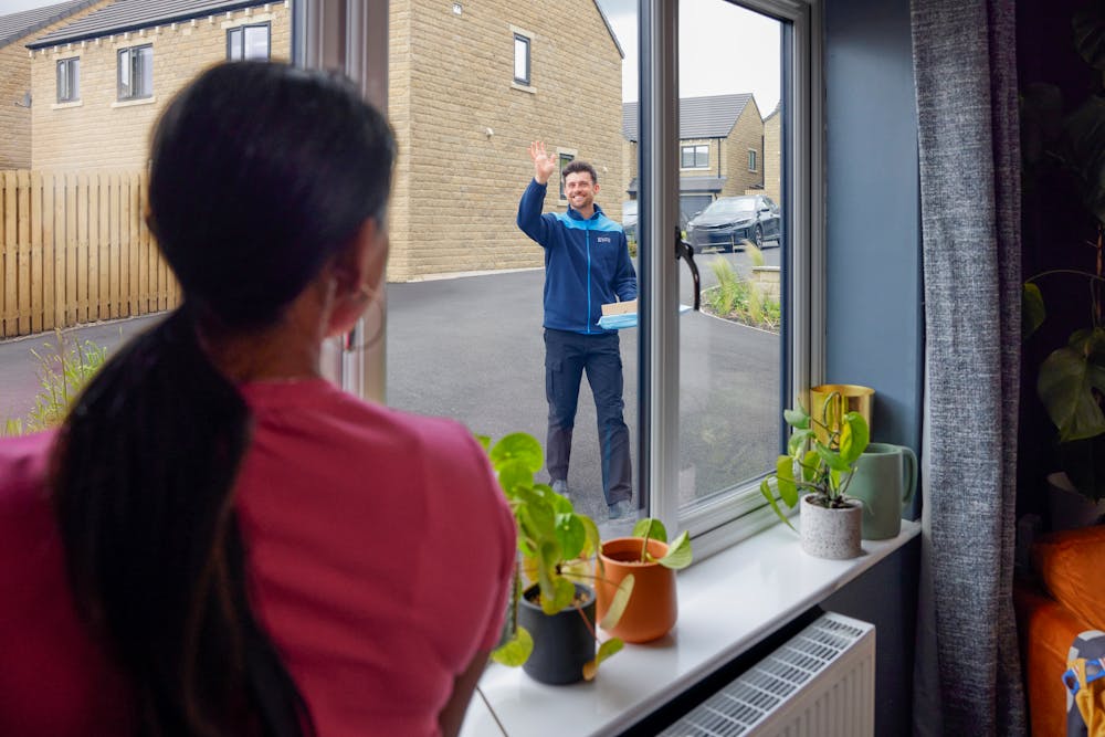 View from the window of a house, an Evri courier is approaching and waving to a woman in the window. He is delivering a couple of parcels to the house.