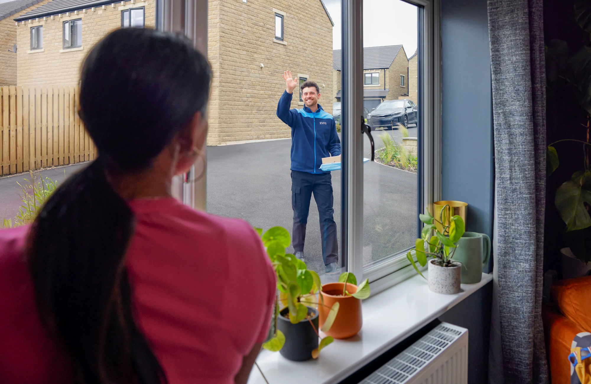 View from the window of a house, an Evri courier is approaching and waving to a woman in the window. He is delivering a couple of parcels to the house.