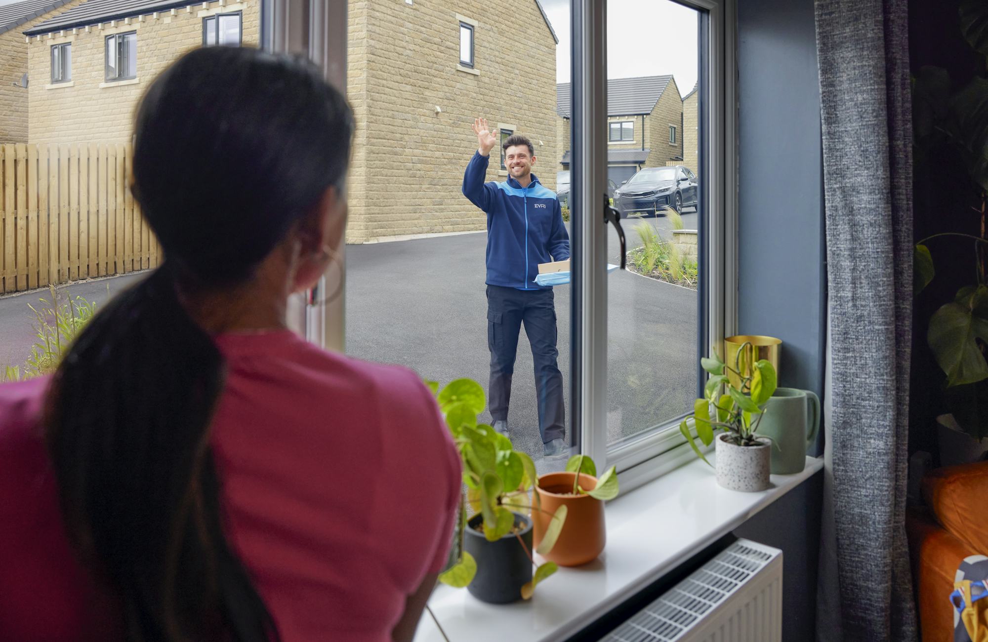 View from the window of a house, an Evri courier is approaching and waving to a woman in the window. He is delivering a couple of parcels to the house.
