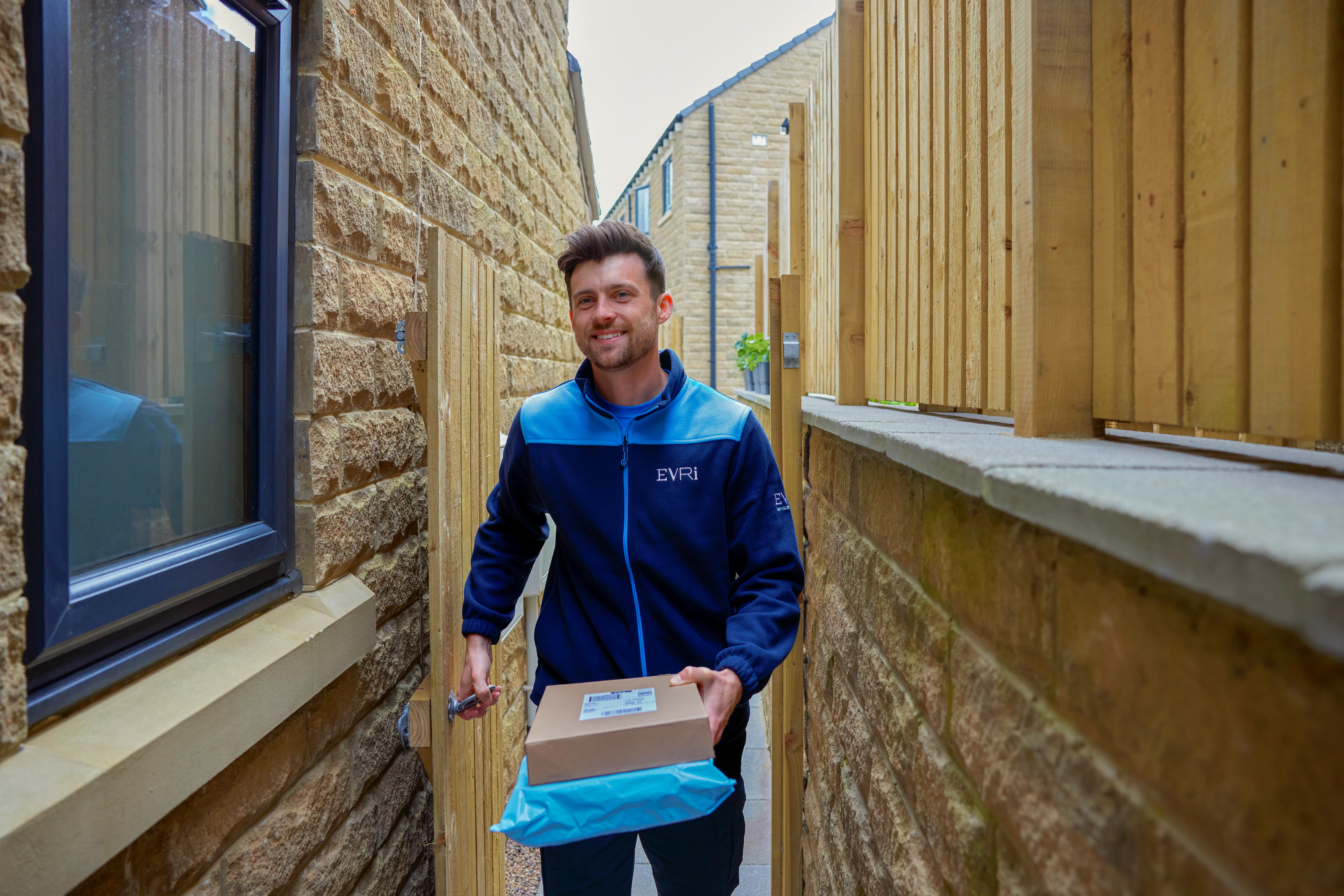 An Evri courier is entering the rear of a house through a side gate. He is carrying two parcels to drop off.