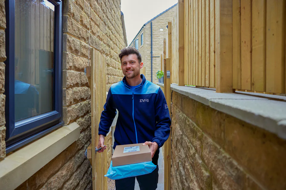 An Evri courier is entering the rear of a house through a side gate. He is carrying two parcels to drop off.