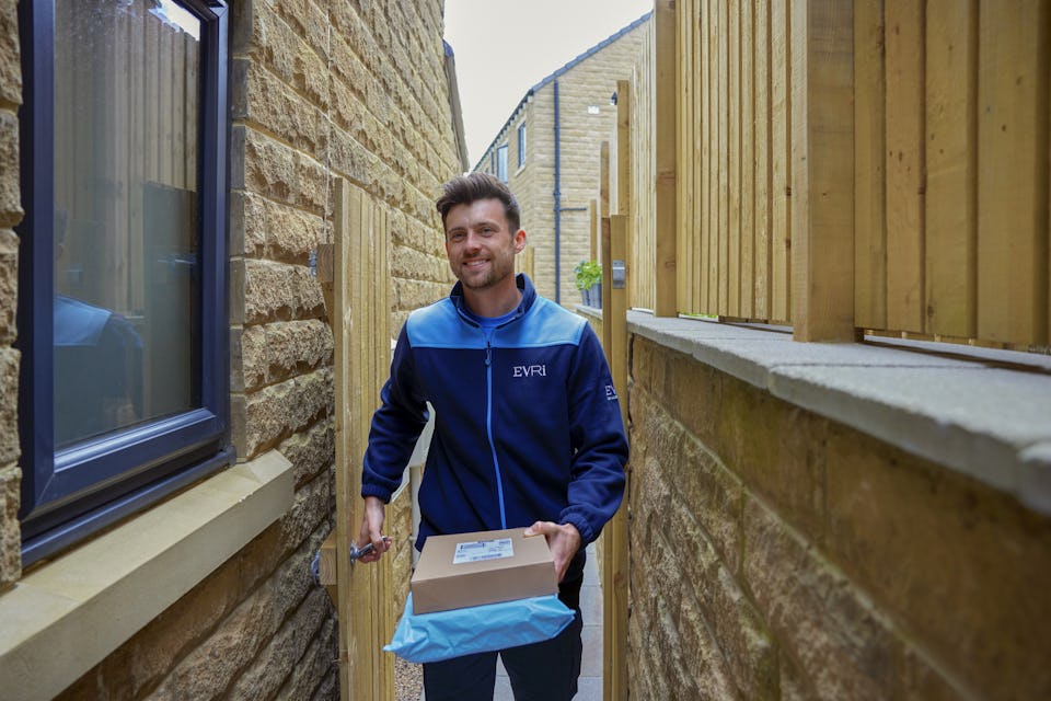 An Evri courier is entering the rear of a house through a side gate. He is carrying two parcels to drop off.