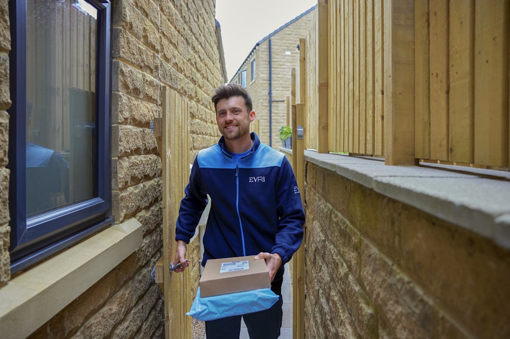 An Evri courier is entering the rear of a house through a side gate. He is carrying two parcels to drop off.