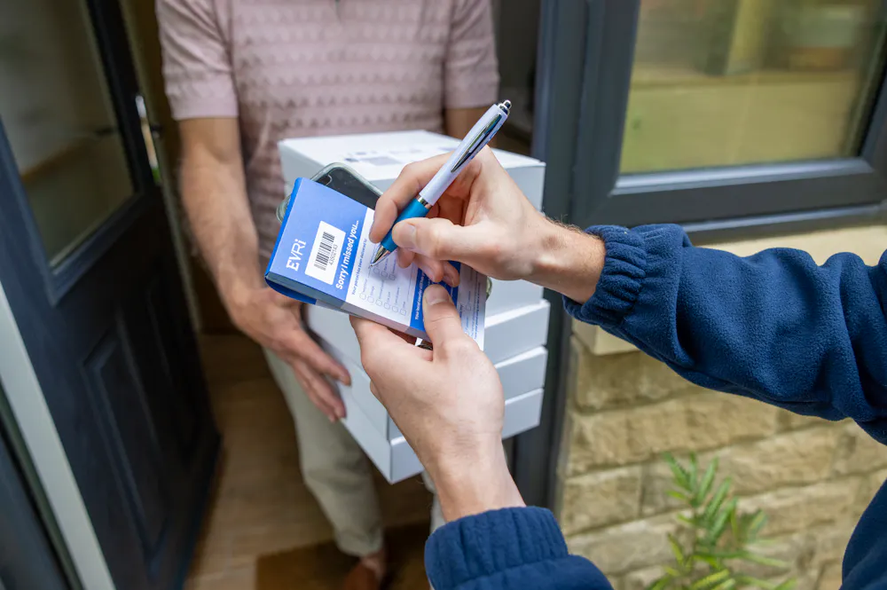 An Evri Courier leaving a stack of parcels with a neighbour due to the recipient not being in. He is taking the name of the neighbour on a blue slip to post to the recipient.