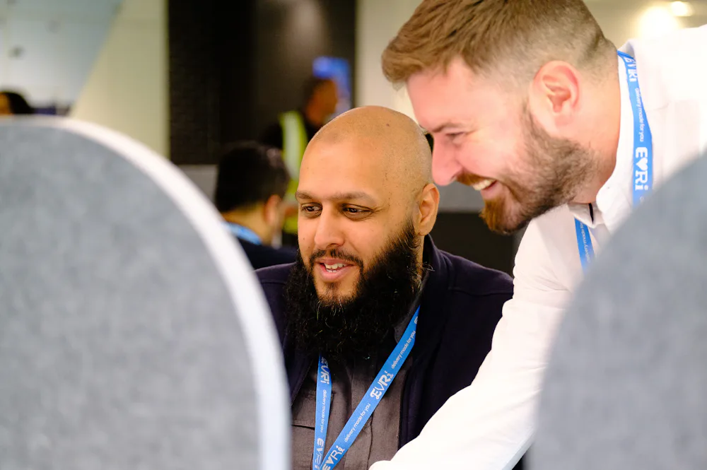 An Evri office worker is sat at his desk whilst another stands over him. They are both looking at their screen in amusement