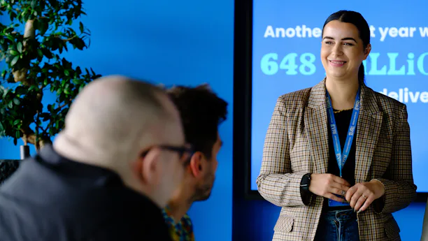 An Evri corporate employee is delivering a presentation in a meeting room. They are stood in front of a large screen displaying some delivery statistics