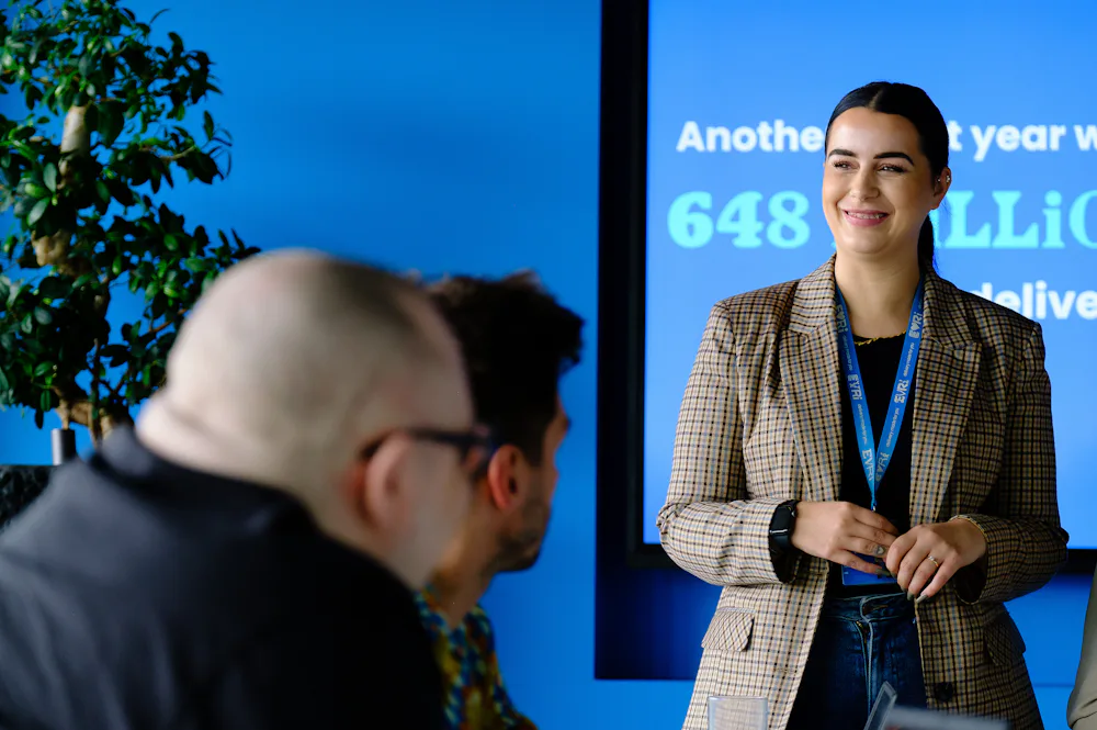 An Evri corporate employee is delivering a presentation in a meeting room. They are stood in front of a large screen displaying some delivery statistics