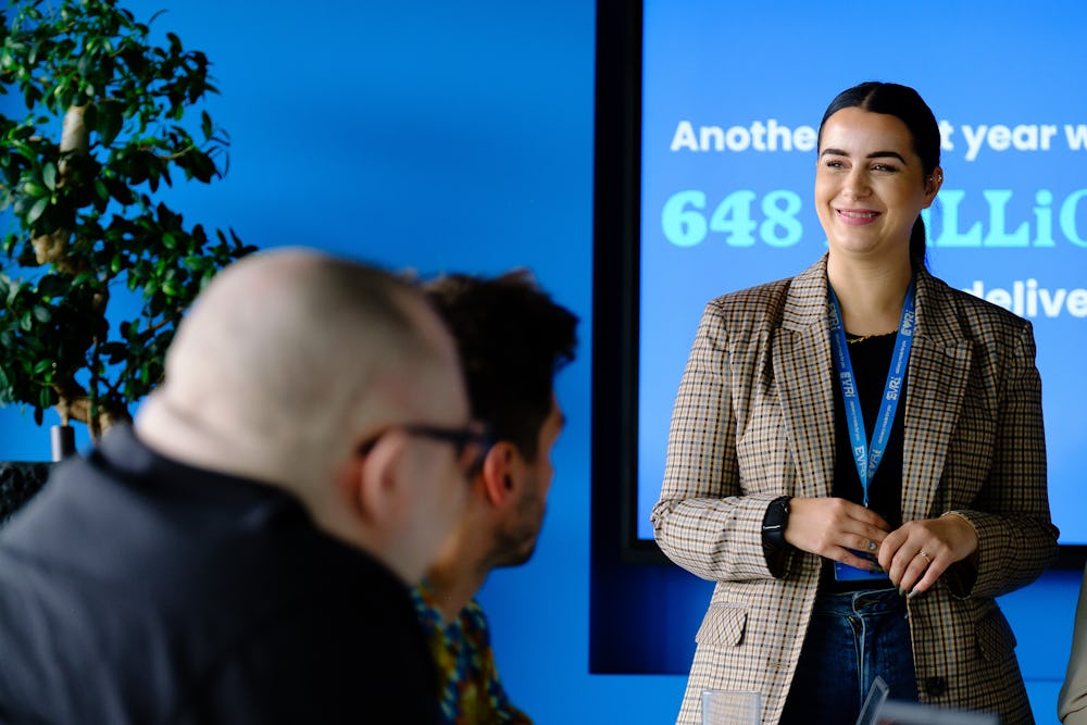 An Evri corporate employee is delivering a presentation in a meeting room. They are stood in front of a large screen displaying some delivery statistics