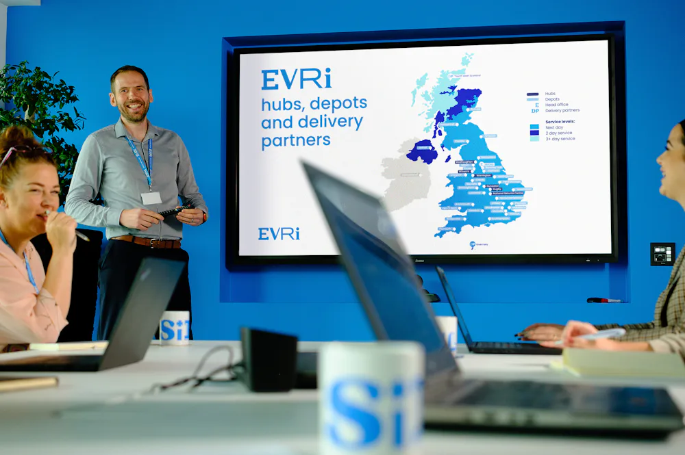 An Evri corporate employee is delivering a presentation in a meeting room. They are stood in front of a large screen displaying some delivery statistics