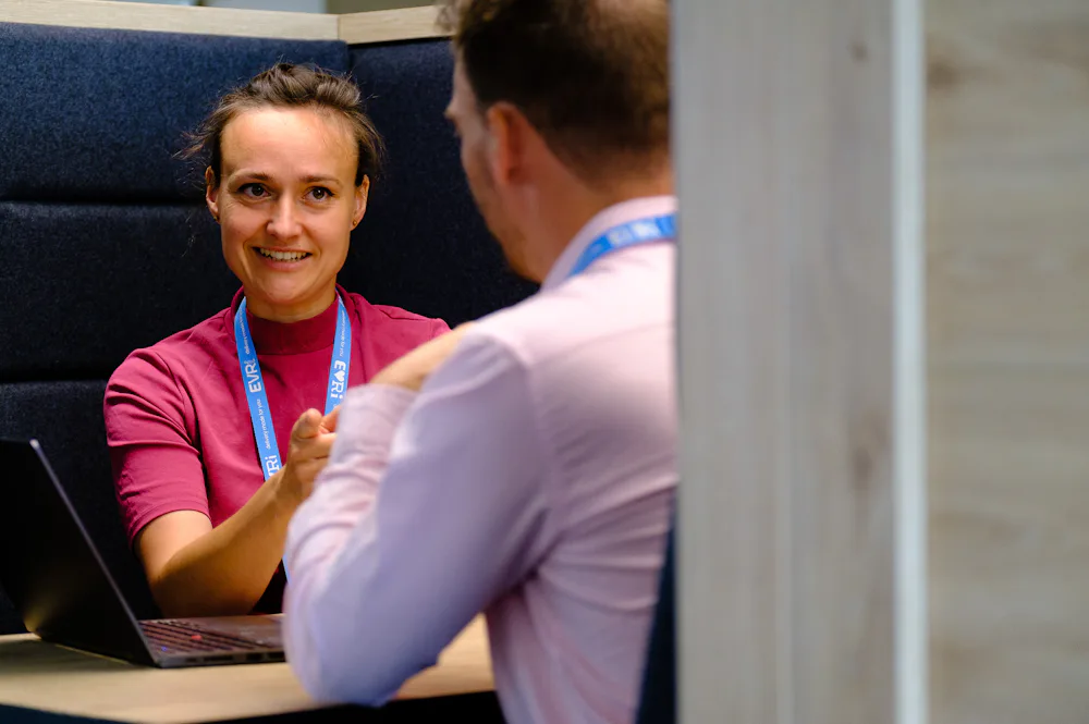 A couple of Evri employees are sat chatting in an office booth with their laptops. They are smiling and pointing at eachother