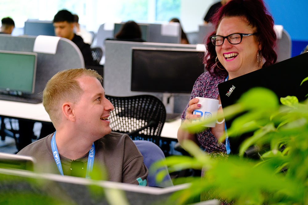 An Evri employee is sat at their desk with another stood beside them. They are laughing at a joke or anecdote.