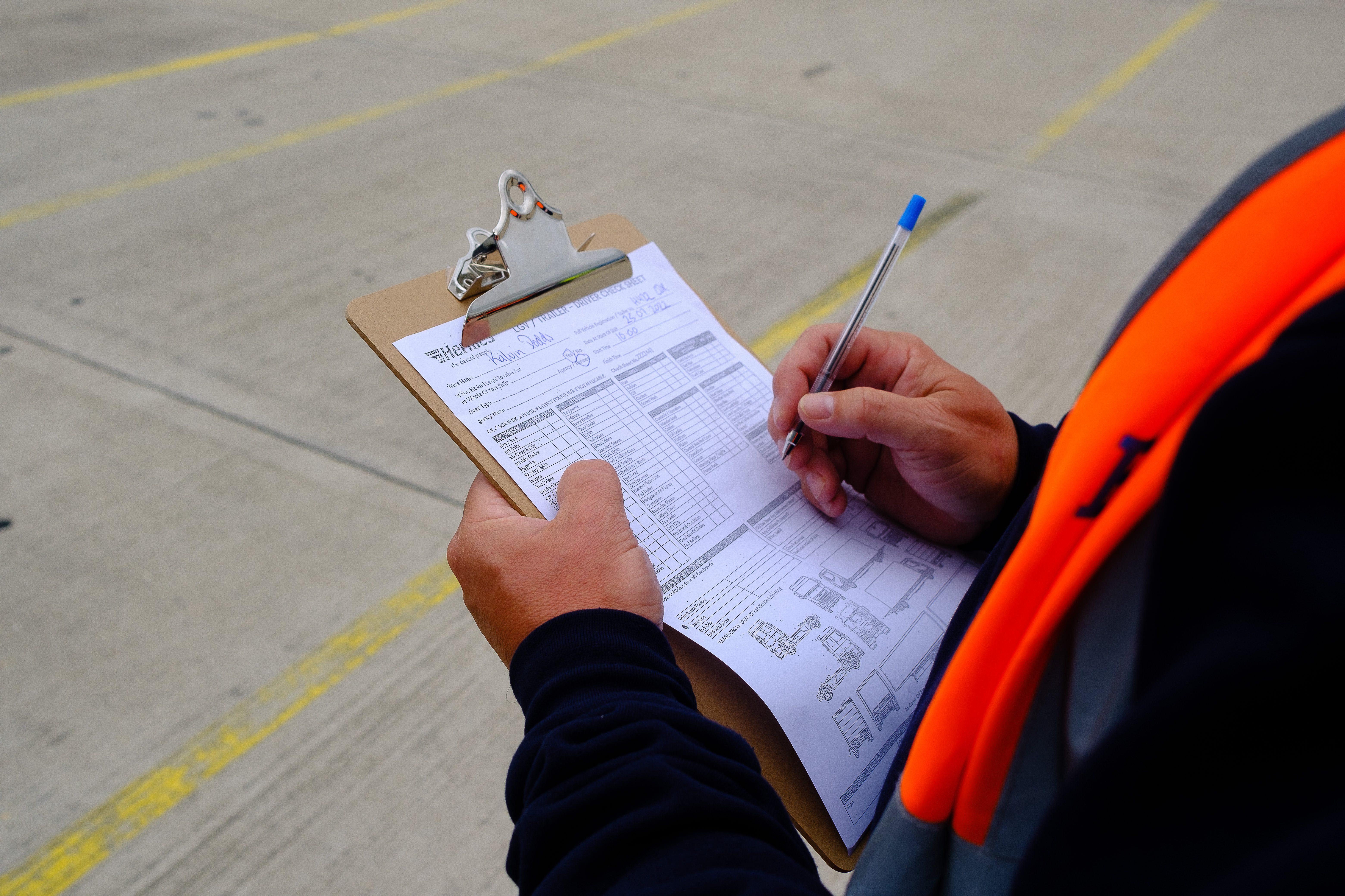 Over the shoulder shot of a man stood outdoors at a depot filling in a checklist attached to a clipboard. He is wearing an orange high vis jacket. 
