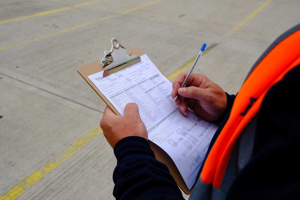 Over the shoulder shot of a man stood outdoors at a depot filling in a checklist attached to a clipboard. He is wearing an orange high vis jacket.