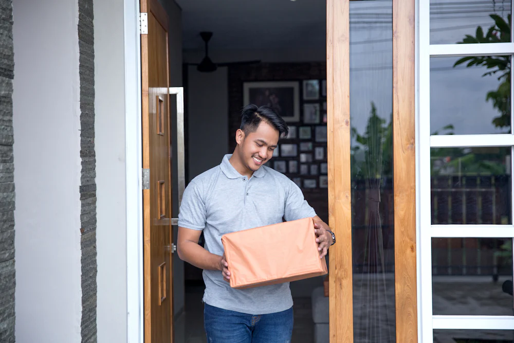 A customer is standing in their doorway holding a large parcel and looking down at it with a smile.