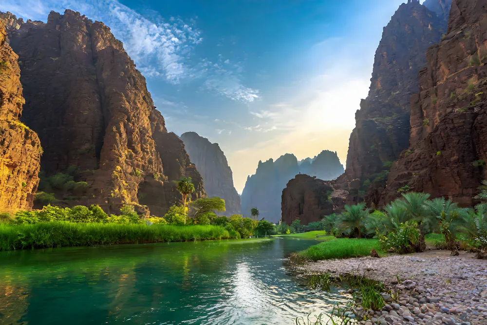 A landscape photo showing a river winding through a rocky-faced mountain range with lots of green shrubbery at the base.
