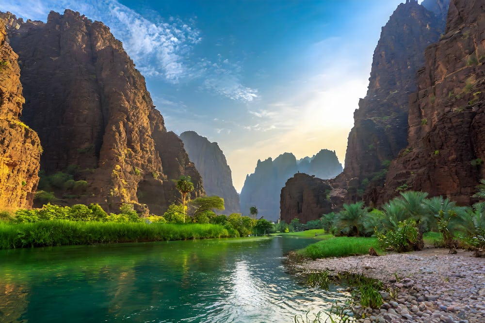 A landscape photo showing a river winding through a rocky-faced mountain range with lots of green shrubbery at the base.