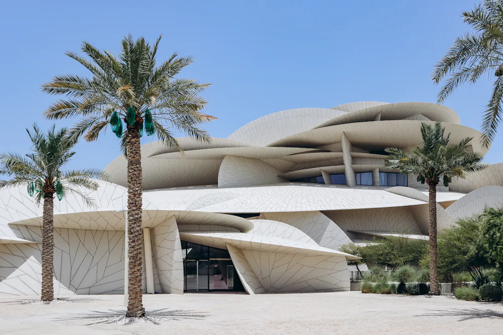 The National Museum of Qatar can be pictured with palm trees in the foreground and clear blue skies. The building is contemporary in design and looks like a desert rose.
