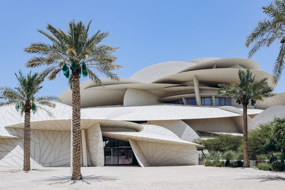 The National Museum of Qatar can be pictured with palm trees in the foreground and clear blue skies. The building is contemporary in design and looks like a desert rose.
