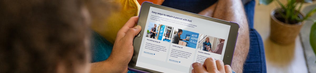 A customer sat on a sofa, holding their tablet and looking at their screen. The display shows the customer is reading about our three easy ways to return a parcel
