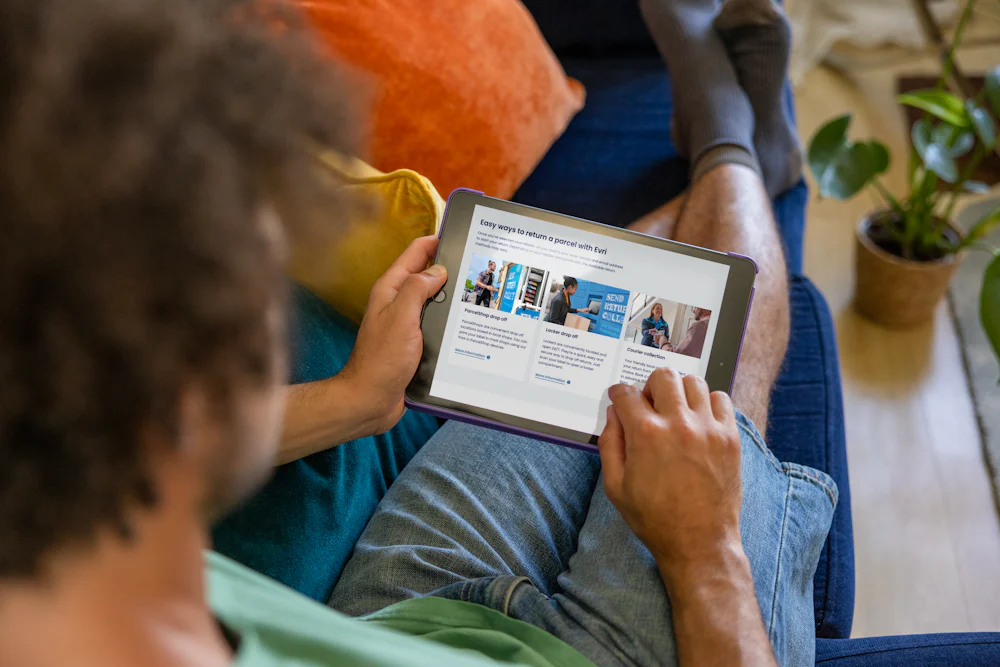 A customer sat on a sofa, holding their tablet and looking at their screen. The display shows the customer is reading about our three easy ways to return a parcel