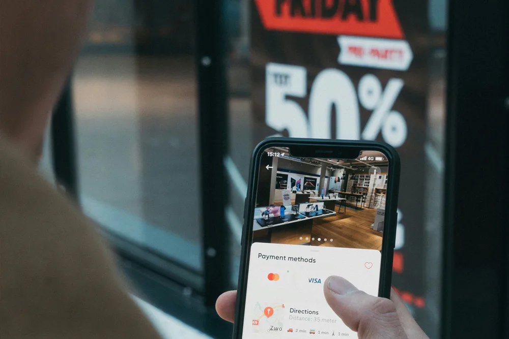 A customer holding their mobile phone with a large Black Friday sale sign in the window of a shop visible in the background