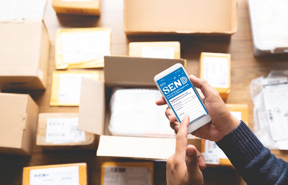 A close-up bird's eye view of a customer holding their mobile phone shows the Evri website home page on display. Lots of parcel boxes can be seen in the background.