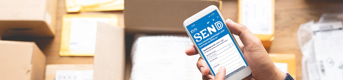 A close-up bird's eye view of a customer holding their mobile phone shows the Evri website home page on display. Lots of parcel boxes can be seen in the background.