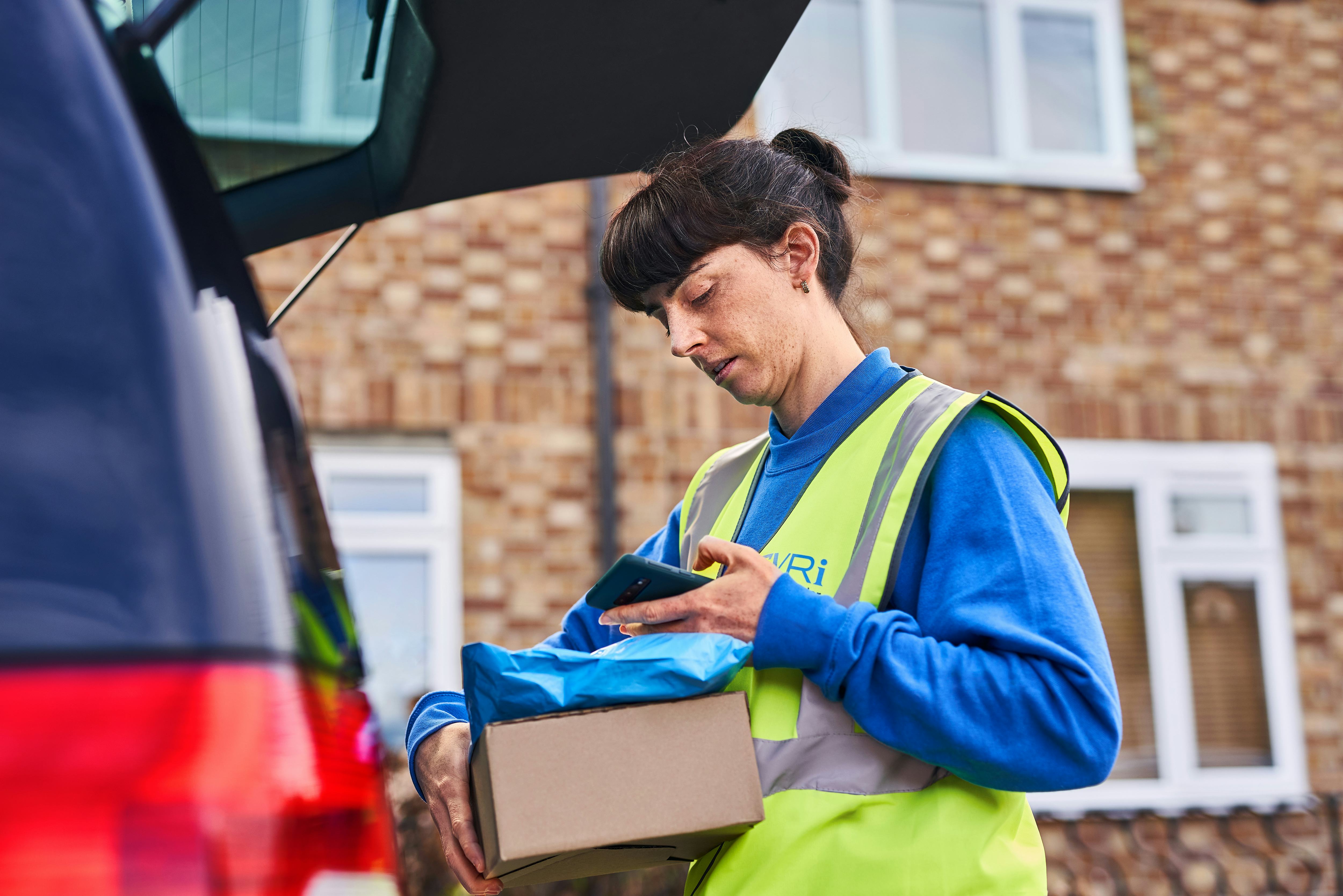 female courier stood under open car boot scanning parcel with her smartphone