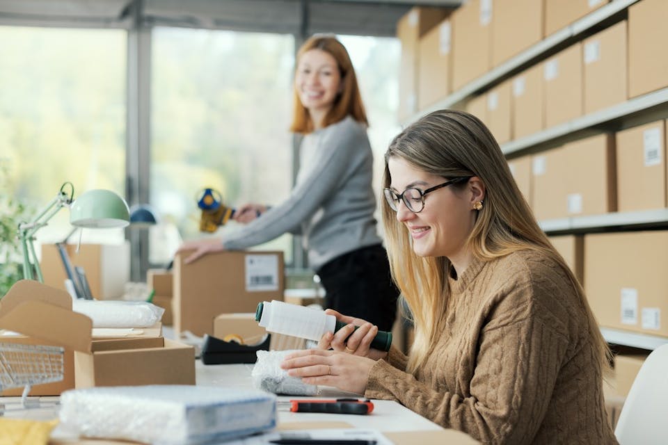 Two female business owners in a stock room seen handling parcels and smiling