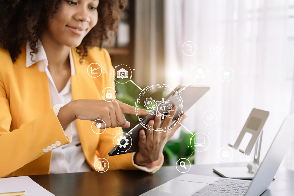 A woman wearing an orange blazer is sitting at a desk in front of an open laptop, holding a tablet and pressing the screen with her other hand. There are digitalized symbols in white overlaying the photo to show she is using AI technology