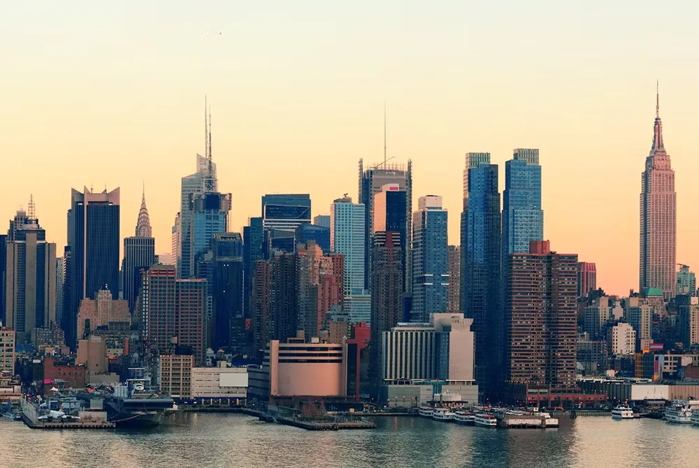 A warm sunset casts golden light over the New York City skyline, highlighting iconic skyscrapers like the Empire State Building as they rise above the waterfront.