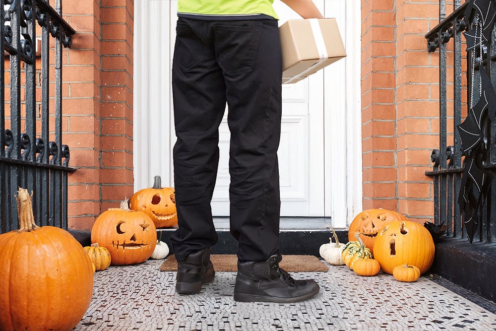 A courier standing on the doorstep, holding a parcel to deliver, with carved pumpkins by their feet.